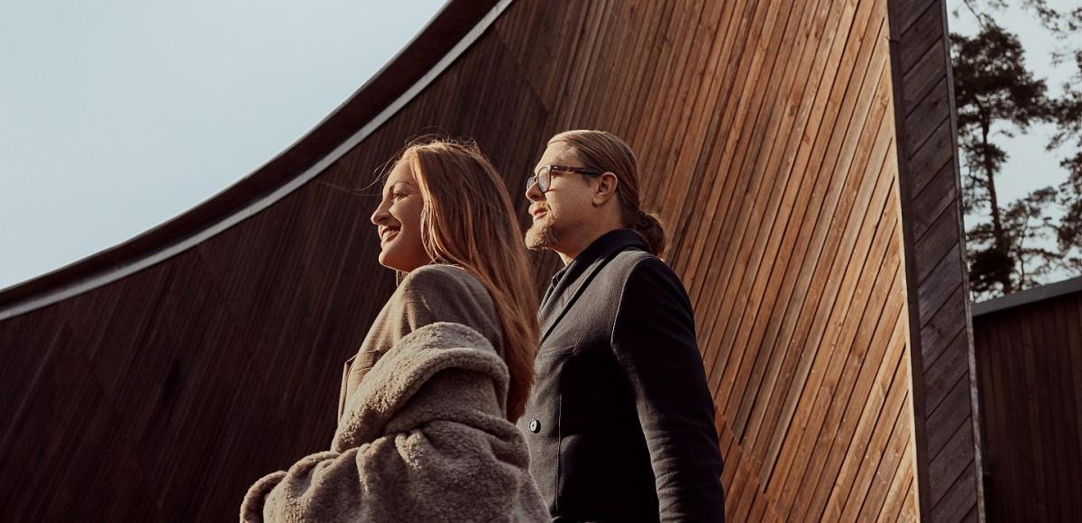 Man with long hair and glasses and a women with a grey coat are looking away to the left